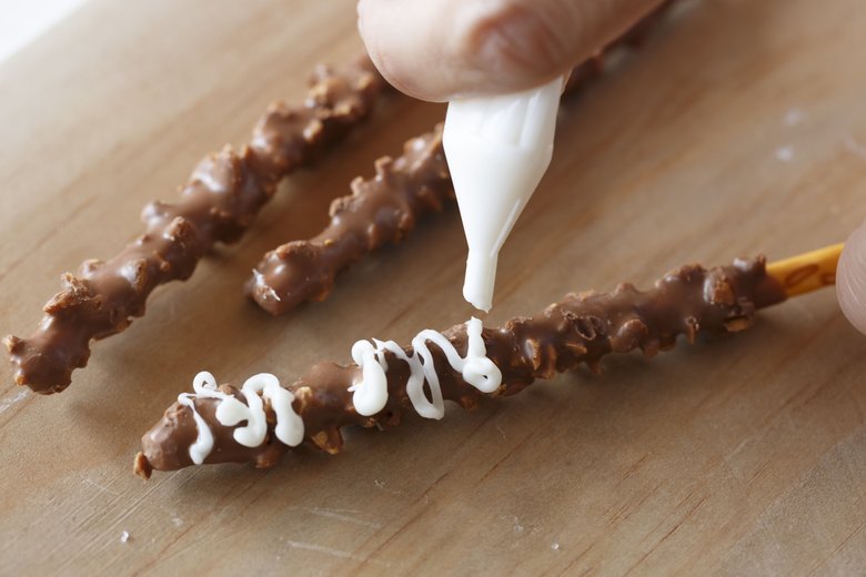 ポッキーやクッキーの表面に、下写真のようにホワイトチョコのチョコペンでジグザグ、ぐるぐる描いていく。ポッキーには、白と茶のチョコペンで目を描いて仕上げる。クリームサンドクッキーは２枚を開いて、開いたクリームの塗られている面にホワイトチョコペンで模様を描く。チョコペンが乾く前に、マシュマロを薄切りにして２つ乗せ、茶のチョコペンで目を描く。
フリーハンドで自由に描いて。子どもにおまかせしてもOK