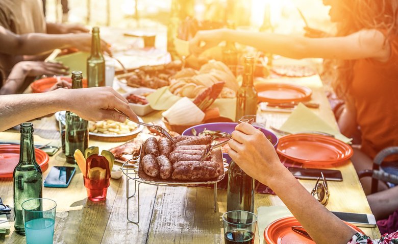 Group of happy friends eating and drinking beers at barbecue dinner at sunset - Adult people having meal together outdoor - Focus on fork sausages - Summer lifestyle, food and friendship concept