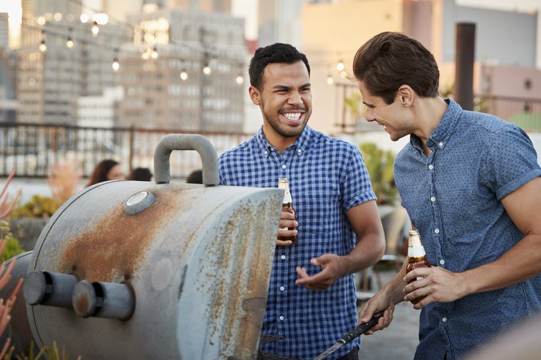 Two Men Cooking Barbecue For Friends Gathered On Rooftop Terrace With City Skyline In Background