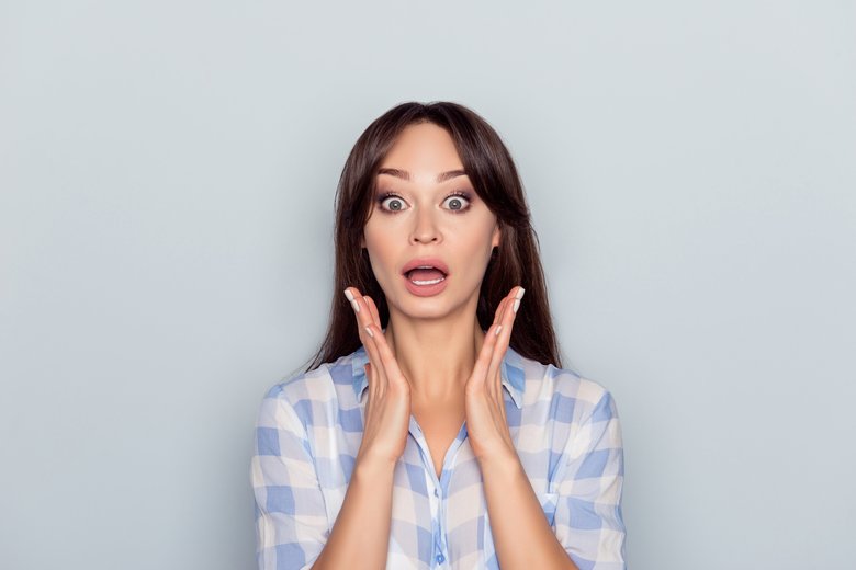 Portrait of pretty, surprised woman with wide opened mouth and eyes in  checkered shirt holding hands near face isolated on gray background