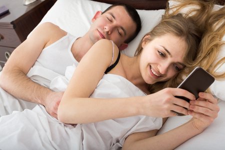 65513134 - positive spanish girl chatting with mobile, man sleeping by in bed