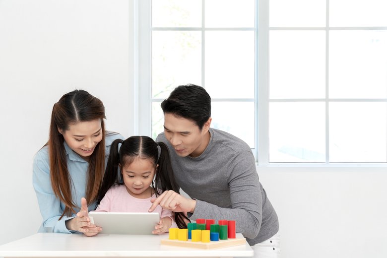 Happy young little asian cute girl watching or playing digital tablet, laptop or mobile with cheerful parents on desk in family time concept in white room with window. Preschool children learning.