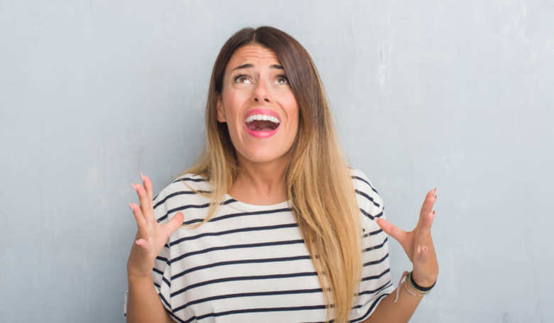 Young adult woman over grunge grey wall wearing navy t-shirt crazy and mad shouting and yelling with aggressive expression and arms raised. Frustration concept.