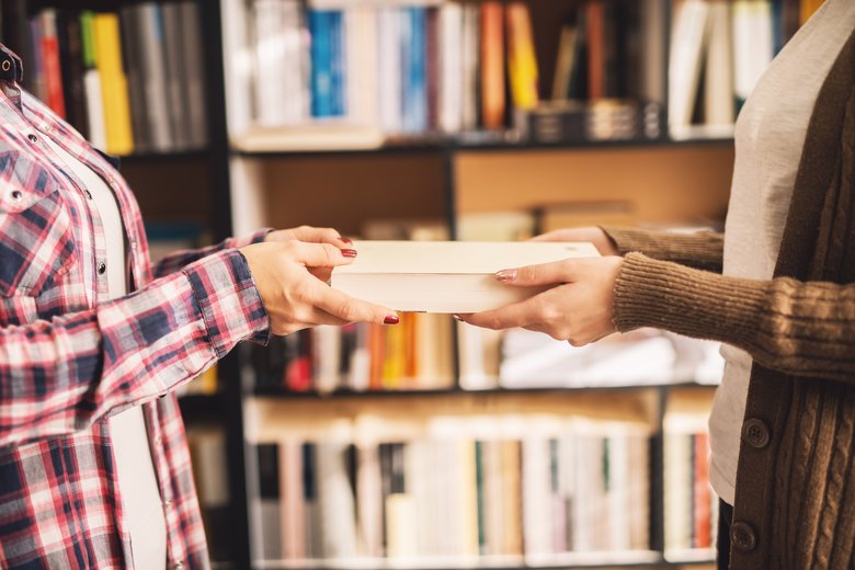 Close up of a girl giving a book to another girl in the library.