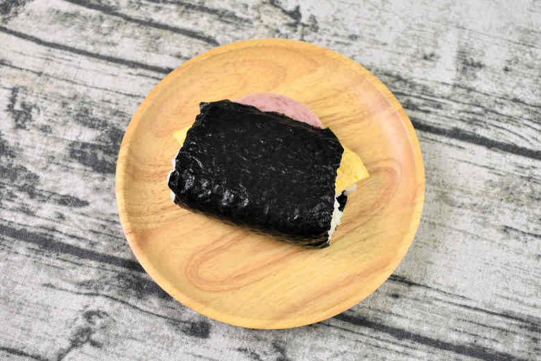 ポップな袋を開けてみると、ご飯にサンドされた具材がお目見え。ピンク色のポークと薄く焼いた玉子焼きが顔を出しています。しかし、パッと見た感じではツナマヨの姿を確認できません。お米の量だけでなく、たっぷりサンドされた具材の食べ応えにも期待が高まりますね。