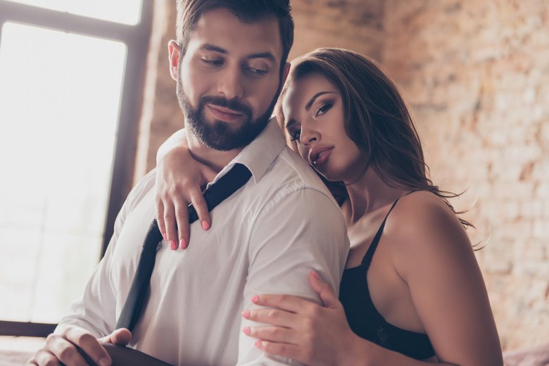 Close up cropped photo of stunning young sexy brunette embracing her lover from the back, he is bearded handsome brunet in a formalwear, smiles. Lady is gorgeous and hot