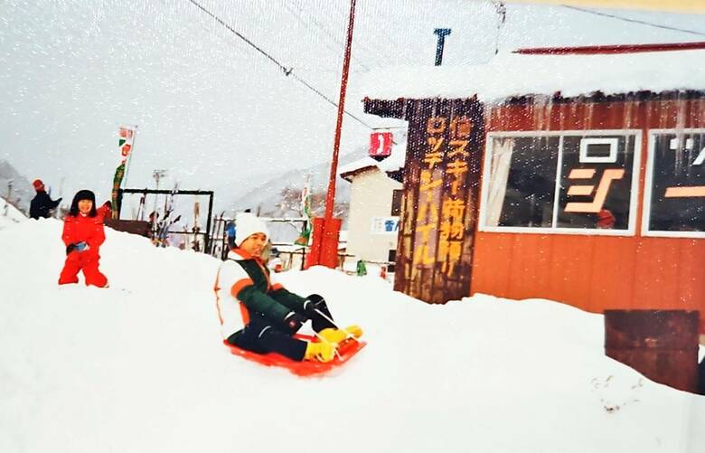 スキー場でソリに乗る鈴木プロデューサーと後ろで笑う麻実子さん